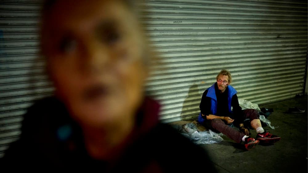Una mujer con trastorno mental se coloca frente a la cámara mientras detrás de ella un hombre pide limosna en Skid Row.