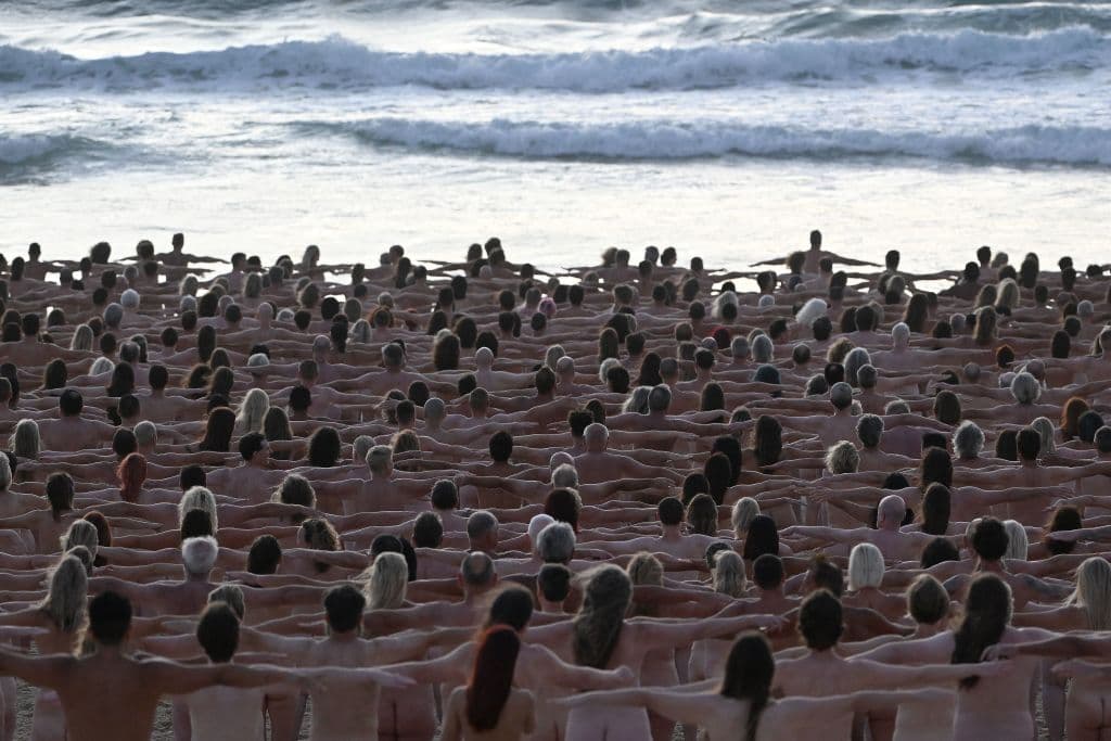En la imagen, miles de voluntarios pozan desnudos con los brazos extendidos en la orilla del mar.