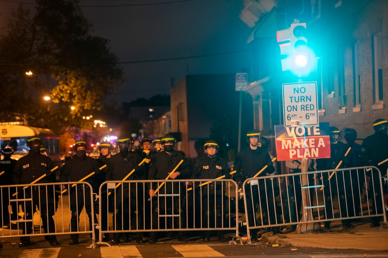 La policía con equipo táctico forma una línea junto a un letrero de "VOTE, HAGA UN PLAN" mientras los manifestantes se reúnen en protesta. El asesinato de Wallace a una semana de las elecciones generales ha colocado a la ciudad en una posición delicada y se espera la presencia de la Guardia Nacional.