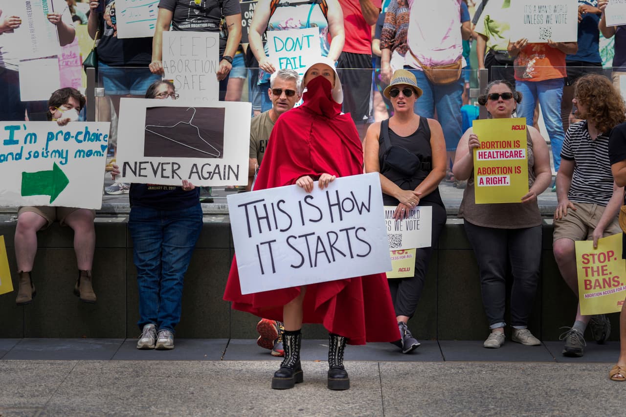 En Indianápolis, Indiana: 
<b>"Así es como empieza".</b>
<br>
<br>La manifestante viste un traje rojo, que refiere a la serie televisiva 
<i>Handsmaid's Tale</i> (El cuento de la criada, en español). La serie muestra una realidad distópica en Gilead, un país fundado tras el fin de EEUU, en el que, en medio de una 'epidemia' de infertilidad, 
<b>las mujeres fértiles son forzadas a tener relaciones bajo un régimen que las trata como propiedades. </b>
<br>
<br>A muchos les preocupa que el fin de Roe v. Wade, que daba base legal "a la privacidad del cuerpo" y permitía a las mujeres tomar sus decisiones reproductivas, abra la puerta a inciertas prohibiciones. 
<br>