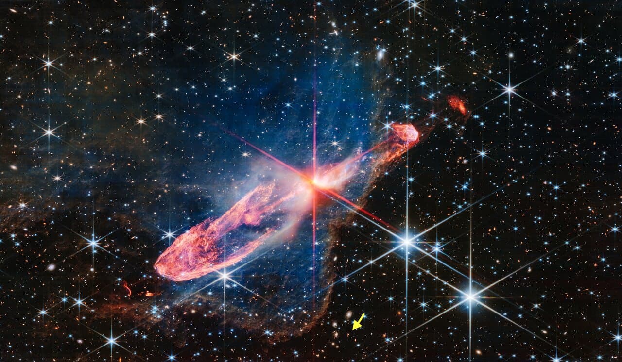 La flecha indica la ubicación del misterioso signo de interrogación en la imagen capturada por el telescopio Webb que muestra un par de estrellas jóvenes muy cercanas conocidas como Herbig-Haro 46/47 rodeadas por un disco de gas y polvo, sobre un fondo de estrellas y galaxias.