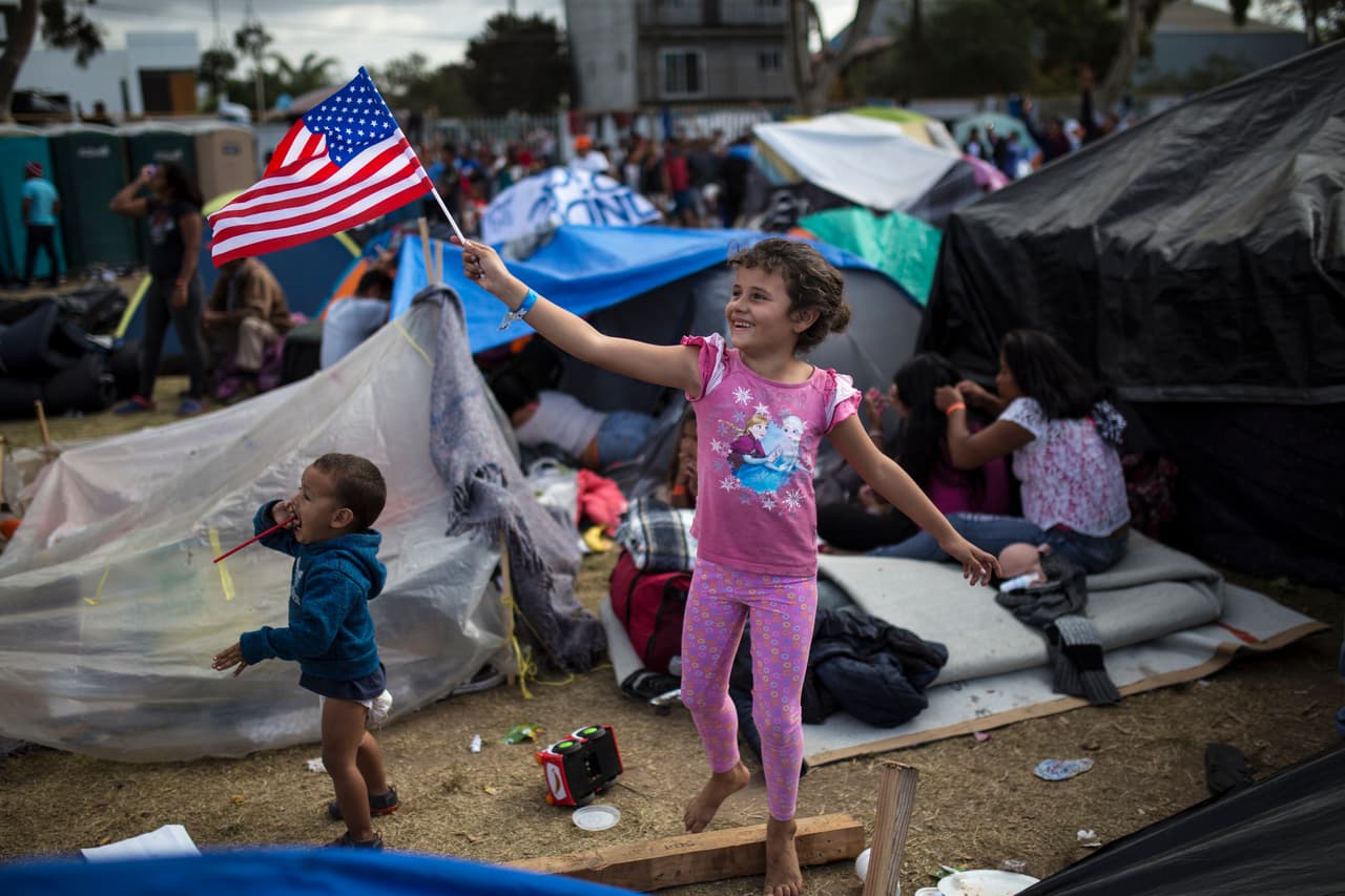 <b>Saludo con la bandera.</b> Más de 5,000 migrantes, muchos de ellos niños, vivieron varias semanas en un refugio con sanitarios portátiles desbordados y un enorme charco de agua sucia que crecía cada vez que alguien tomaba una ducha. 
<a href="https://www.univision.com/noticias/inmigracion/aguantar-o-volver-las-condiciones-infrahumanas-que-soportan-los-migrantes-en-el-refugio-colapsado-de-tijuana-fotos-fotos">Las condiciones deplorables de la Unidad Deportiva Benito Juárez, donde fueron alojados la mayoría de los centroamericanos, impulsaron a algunos a regresar a sus países o a buscar ingresar a EEUU ilegalmente.</a> En la fotografía, tomada el 24 de noviembre de 2018, la pequeña hondureña Génesis Belén Mejía Flores saluda a un helicóptero estadounidense que volaba cerca del albergue, ubicado al norte de Tijuana.