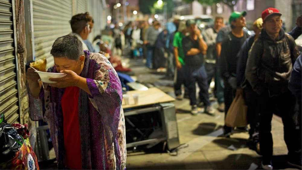 Una mujer come un caldo de pollo mientras otros indigentes hacen fila para recibir alimento.