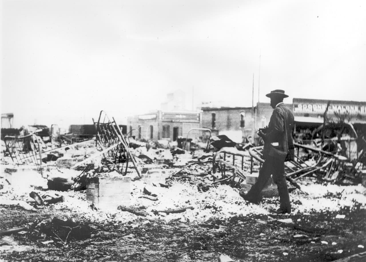 <b>Cenizas y escombros tras la matanza.</b>
<br>
<br>En la fotografía un hombre afroamericano con una cámara junto a los esqueletos de las camas de hierro sobre las cenizas de una cuadra devastada.
<br>