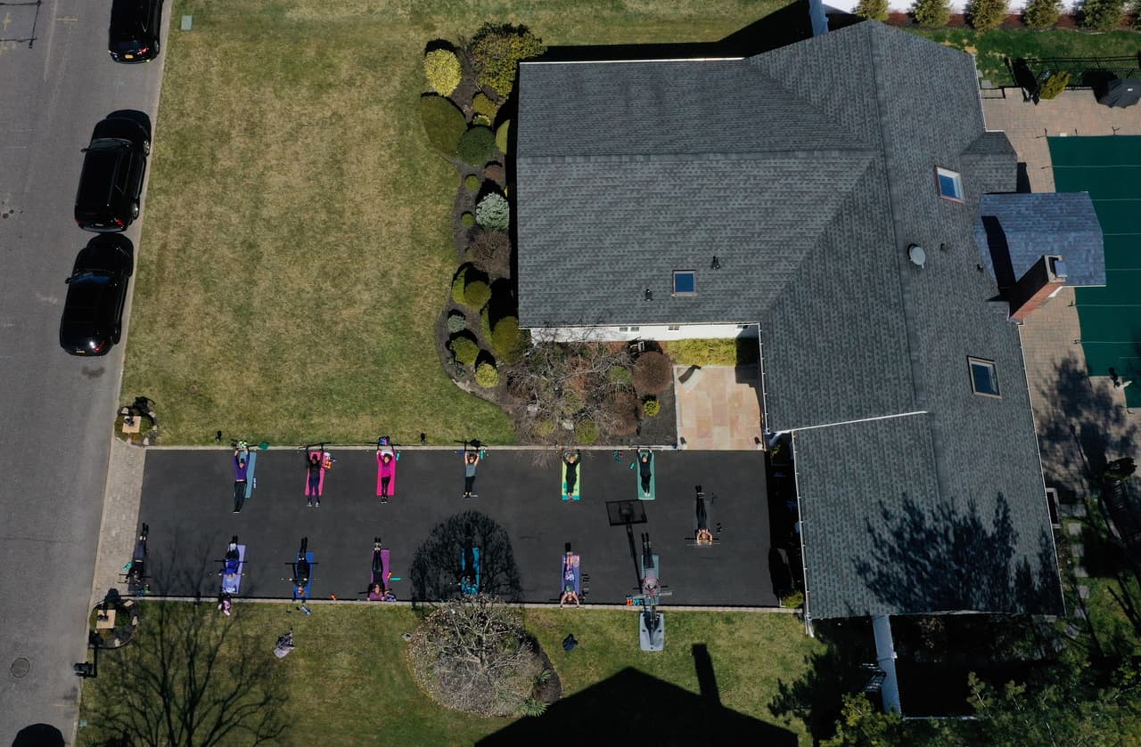 <b>Acondicionamiento físico con seis pies de separación.</b> Jamie Benedik, instructora física de West Islip, Nueva York, imparte una clase en la entrada de su casa, el 26 de marzo. Ella es instructora en tres gimnasios en su área pero están cerrados, por eso convoca a sus alumnos la entrada de su estacionamiento.