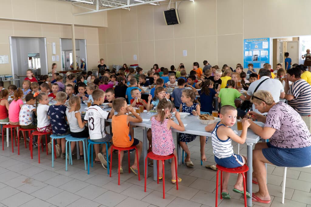 Niños ucranianos procedentes de diferentes orfanatos de la región de Donetsk, durante una comida en un campamento en Zolotaya Kosa, en el suroeste de Rusia. La defensora de los derechos de los niños rusos Maria Lvova-Belova, sobre quien recaen 
<a href="https://www.univision.com/noticias/mundo/las-sanciones-contra-putin-ucrania">sanciones de varios países occidentales</a>, ha dicho que estos niños necesitan la ayuda de Rusia para superar el trauma que les ha hecho 
<b>dormir mal, llorar por la noche y dibujar sótanos y refugios antibombas</b>. Reconoció que al principio, un grupo de 30 niños traídos a Rusia desde los sótanos de Mariupol 
<b>cantaban desafiantemente el himno nacional ucraniano y gritaban "¡Gloria a Ucrania!"</b>. Pero ahora, dijo, sus críticas se han "transformado en amor por Rusia", y ella misma ha acogido a uno, un adolescente.