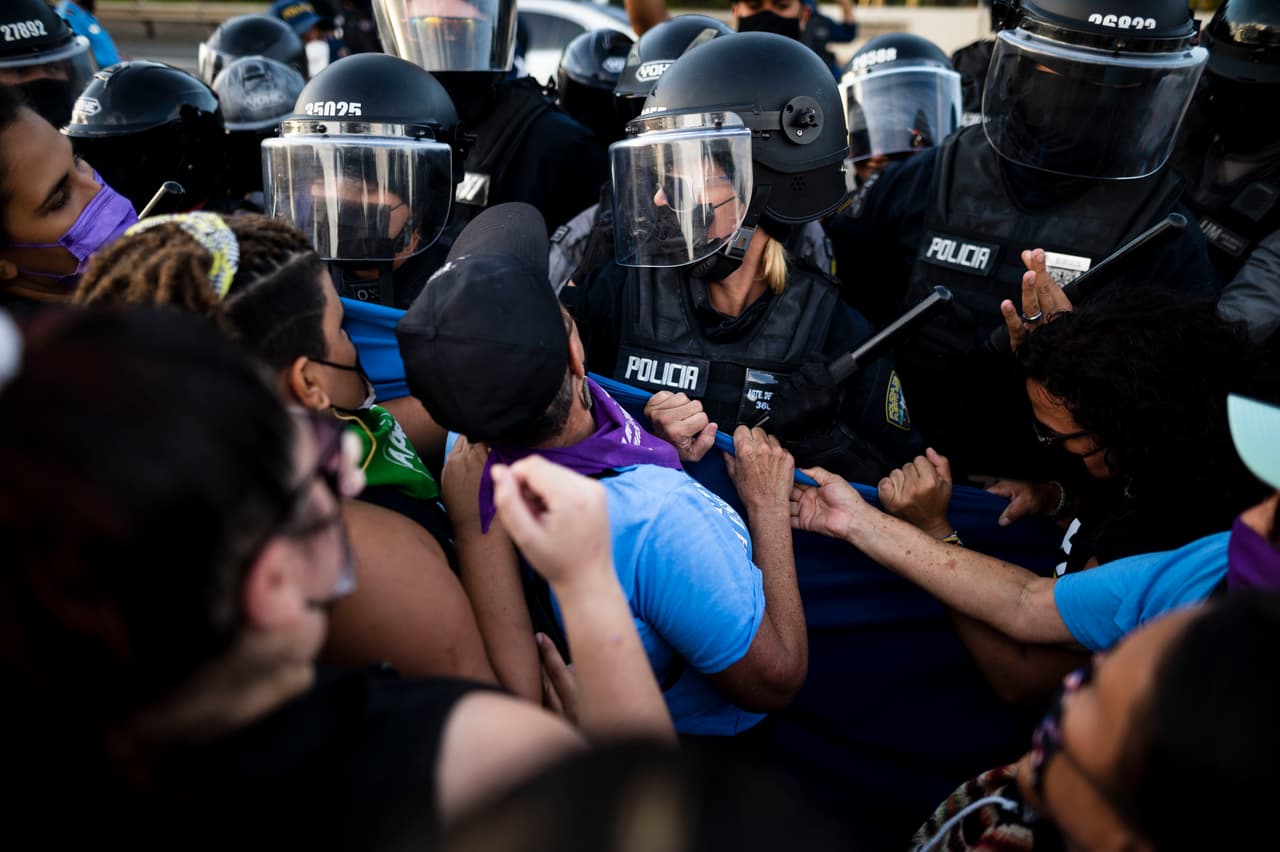Un grupo de manifestantes feministas se enfrenta a la policía mientras bloquean el tráfico en una autopista, protestando por la privatización de los servicios públicos de energía y exigiendo al gobierno rescindir el contrato con LUMA Energy.
<br>