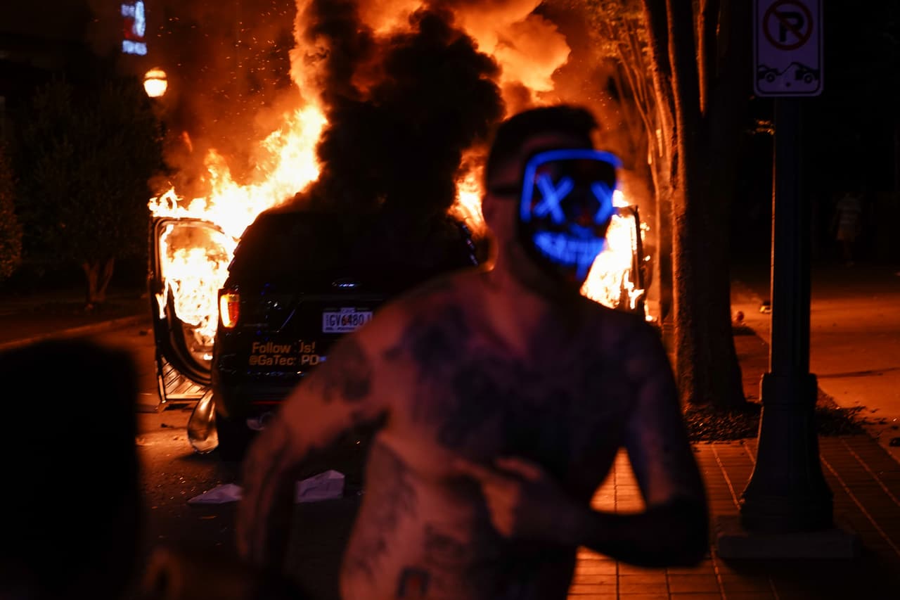 Un hombre con una máscara de 'Purga' corre delante de un coche de policía en llamas durante una protesta el 29 de mayo de 2020 en Atlanta, Georgia.