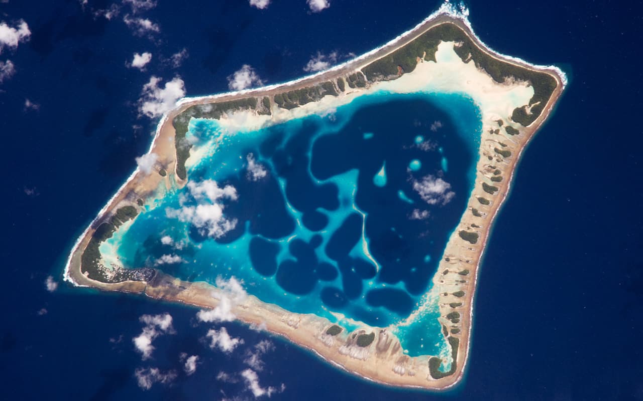 <b>Atolón de Atafu, Tokelau. </b>Con aproximadamente ocho kilómetros de ancho, el atolón de Atafu es el más pequeño de tres atolones que comprende el grupo de islas Tokelau, en el sur del Océano Pacífico. Es actualmente un territorio de Nueva Zelanda aunque se han celebrado referéndums sobre la independencia de las islas. El asentamiento principal en Atafu es un pueblo (casi en el extremo izquierdo, visibles puntos grises) que se ve en esta fotografía de un astronauta. La típica forma de anillo del atolón es el resultado de la acumulación de arrecifes de coral alrededor de una antigua isla volcánica. Con el tiempo geológico, el volcán central se ha hundido 
<br>