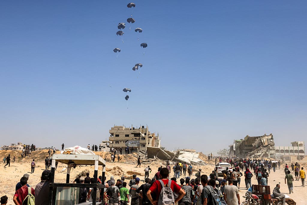 Varias personas se reúnen mientras un avión de transporte militar Hércules C-130 deja caer ayuda humanitaria sobre el norte de la Franja de Gaza el 27 de julio de 2025. Dos aviones jordanos y uno emiratí lanzaron 25 toneladas de ayuda humanitaria sobre la Franja de Gaza, según informó el 27 de julio la televisión estatal jordana.