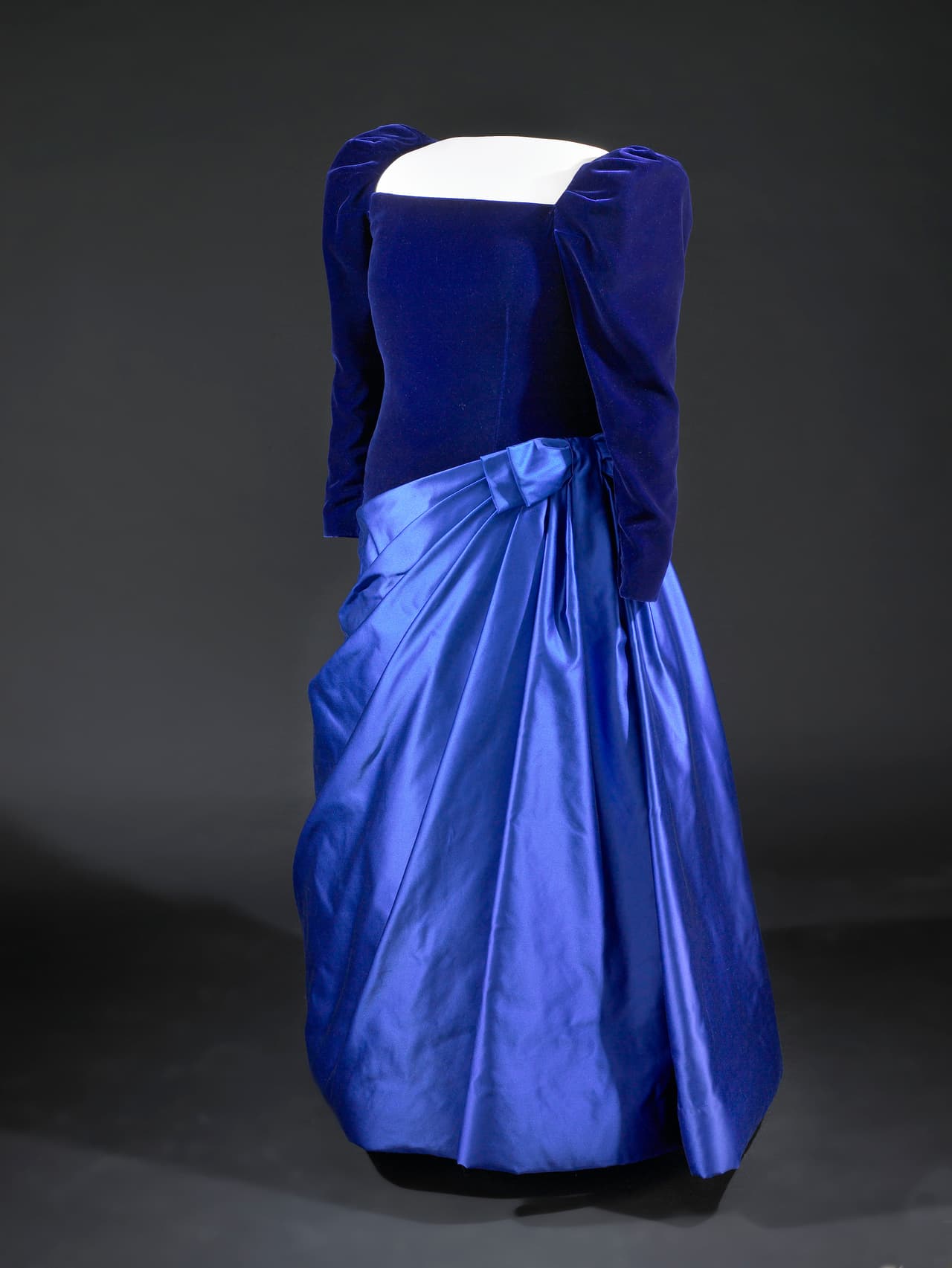 Este vestido creado por 
<b>Arnold Scassi </b>es un perfecto resumen del estilo de la década de los 80. El terciopelo era el rey, así como las hombreras y mangas abombadas. Su accesorio, unas perlas que se convertirían en su marca registrada. Su clutch sería diseñado por Judith Leiber, que sería una constante entre las mujeres del poder incluso unas cuantas décadas después.