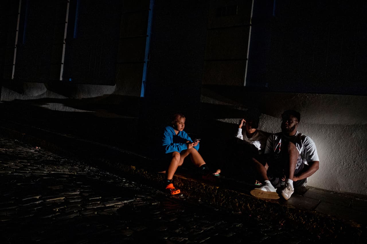 La gente se sienta en la acera de una calle que quedó a oscuras por un corte de energía tras el incendio en una subestación eléctrica en Puerto Rico.
