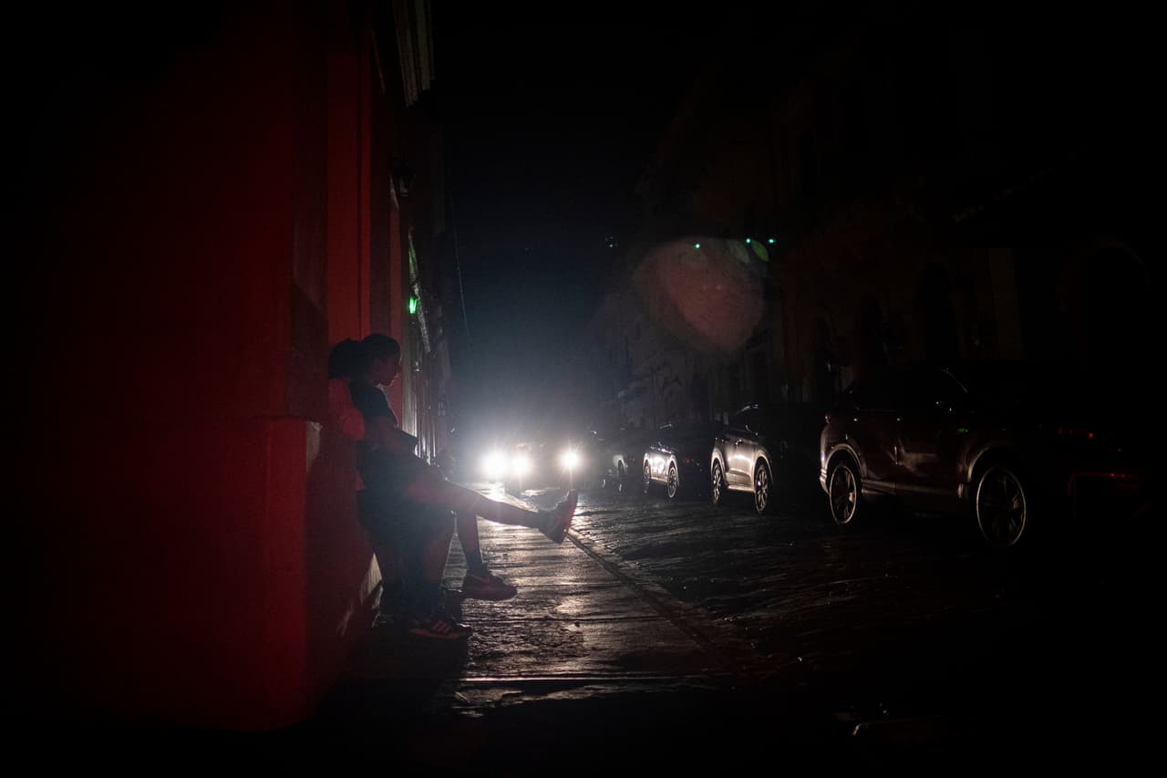 La gente se sienta en la acera de una calle que quedó a oscuras por un corte de energía debido a un ciberataque en el Viejo San Juan.