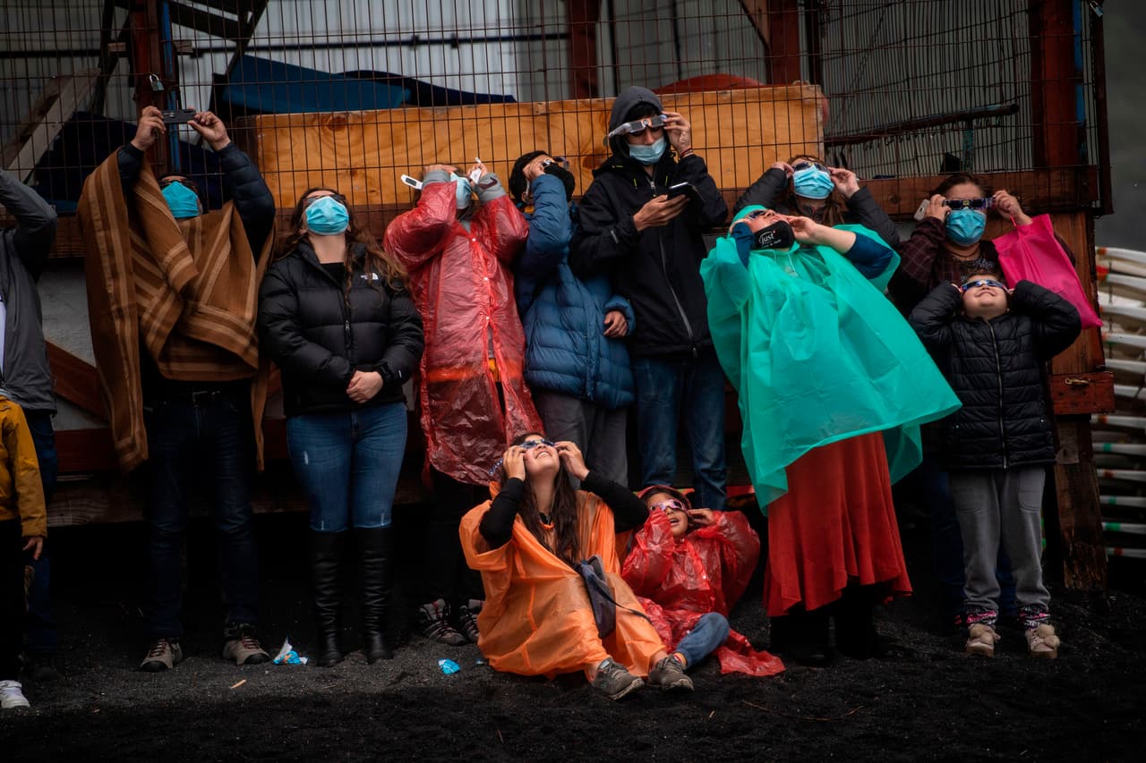 Personas observan el eclipse total de sol en las orillas del lago Villarrica, Chile, utilizando máscaras para evitar la propagación del coronavirus y ropa gruesa para protegerse de la fuerte lluvia que azotó la región horas antes del suceso de este lunes 14 de diciembre. 
<br>
<br>Este año el eclipse tiene lugar en plena pandemia del covid-19, que ha dejado en Chile más de 560,000 contagios y que en las últimas semanas ha dado señales de una segunda ola, por lo que las autoridades insistieron en que se cumplieran las restricciones sanitarias y evitaran aglomeraciones durante el evento.