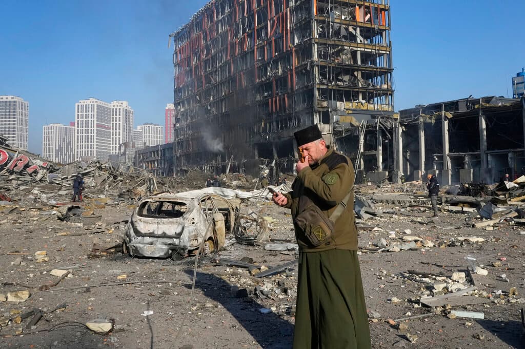 Un sacerdote reza frente a las ruinas del centro comercial tras el ataque ruso, que también 
<b>destruyó todo lo que había alrededor</b>, edificaciones y vehículos.